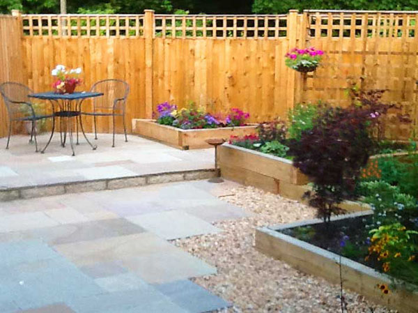 Indian sandstone paving sparkles beside timber raised beds. Bistro table and chairs at far end on sunny raised terrace